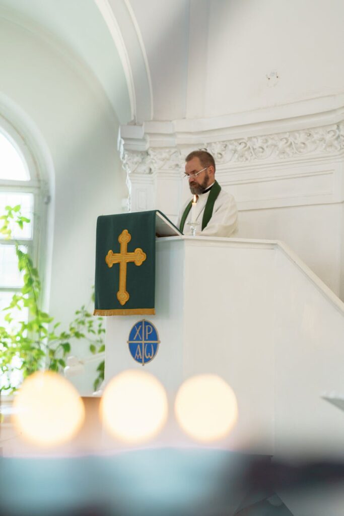 A pastor delivering a sermon inside a bright church with greenery and warm lighting.